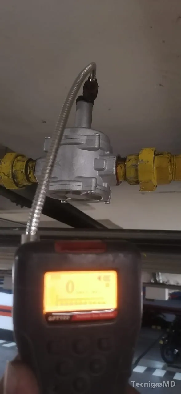 Explosímetro con sonda flexible acercado a un regulador de gas natural en tubería amarilla bajo el techo de un parqueadero