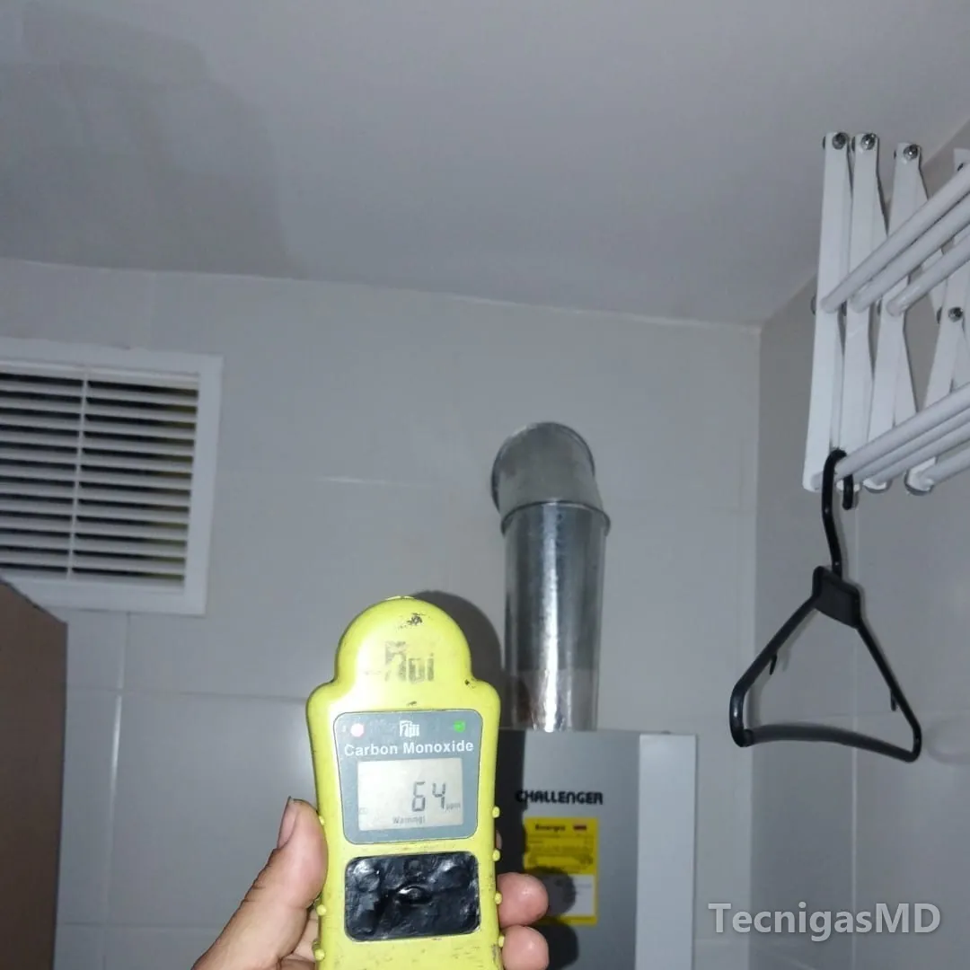 Mano sostiene un detector de monóxido de carbono frente a un calentador a gas Challenger con ducto de evacuación metálico