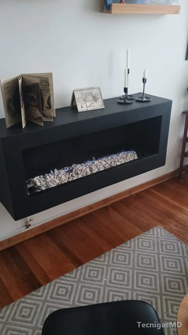 Chimenea a gas lineal de marco negro suspendida en el muro de una sala, con llama azul baja sobre piedras decorativas
